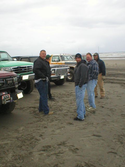 1978 Ford Bronco - Several trucks are parked on a beach as a group of men stand around.