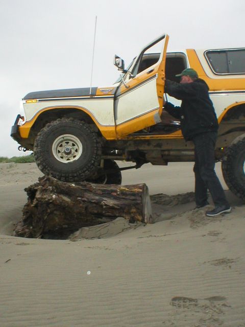1978 Ford Bronco - A man enters a yellow and white 1978 Ford Bronco parked on a sandy beach, with the front tire resting on a large log.