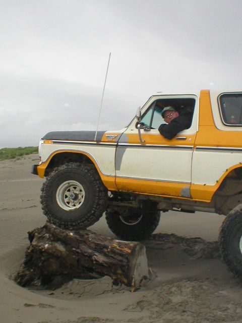 1978 Ford Bronco - A driver navigates a lifted Ford Bronco over a large log on a sandy beach under an overcast sky.