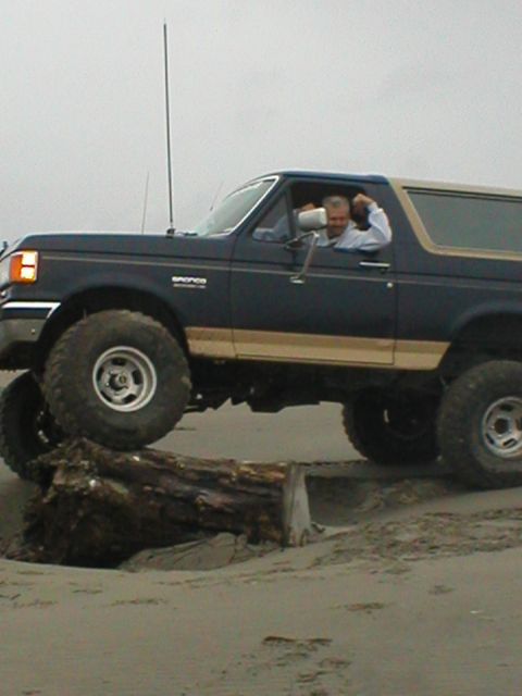 1978 Ford Bronco - A man drives a dark blue SUV with a tan top over a log on a sandy beach, demonstrating its off-road capability.