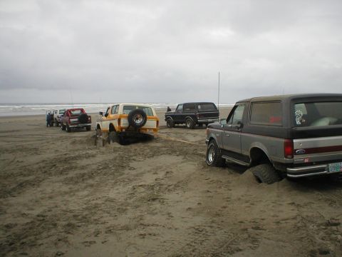 1978 Ford Bronco - Multiple Ford Broncos struggle in the sand on a beach, with one vehicle stuck deep and another assisting.
