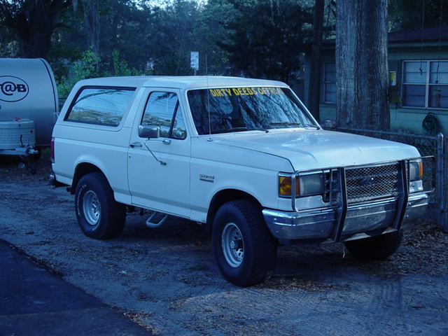 White Ford Bronco with brush guard and 'Dirty Deeds Offrd' windshield banner, 3/4 front view on dirt driveway