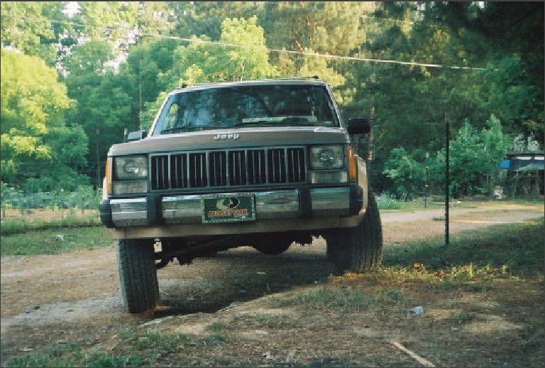 Front view of tan Jeep Cherokee XJ with Mossy Oak front plate on dirt driveway among trees