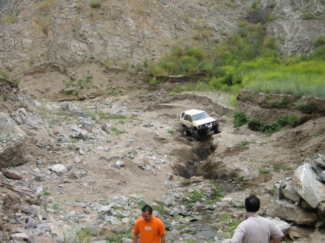 Lifted white SUV crawling down a rutted dirt gully as two men watch