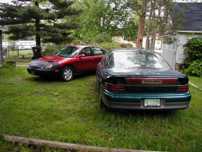 1997 Ford Taurus - Two sedans are parked in a yard, one is a red 1997 Ford Taurus with a car cover.