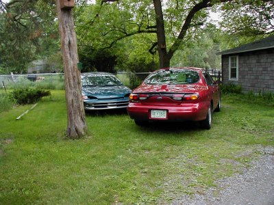 1997 Ford Taurus - Two sedans are parked in a grassy yard next to a house.