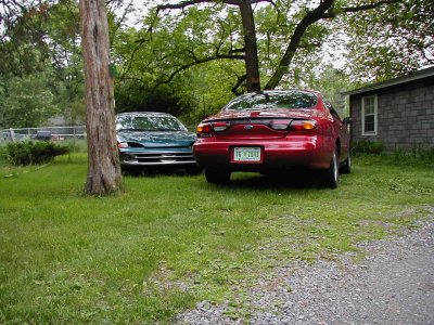 1997 Ford Taurus - Two sedans are parked in a yard next to a building, one red and one green.