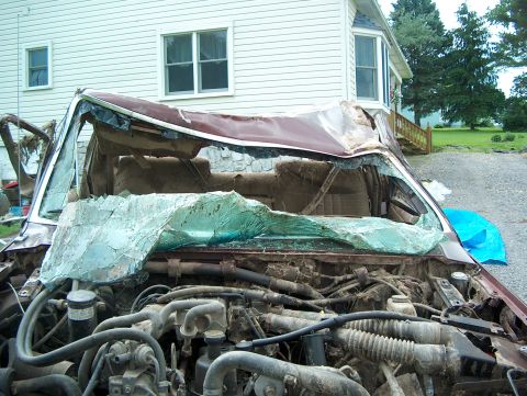 1990 Ford F-150 - A severely damaged 1990 Ford F-150 truck sits in front of a house, its windshield shattered and roof caved in.
