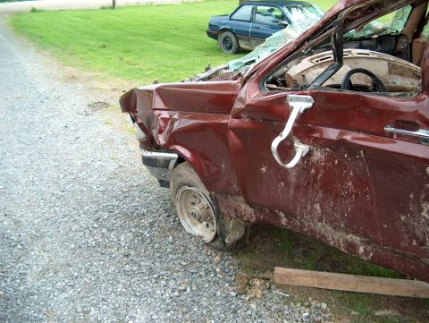 1990 Ford F-150 - A severely damaged maroon pickup truck sits on a gravel lot, with another vehicle visible in the background.