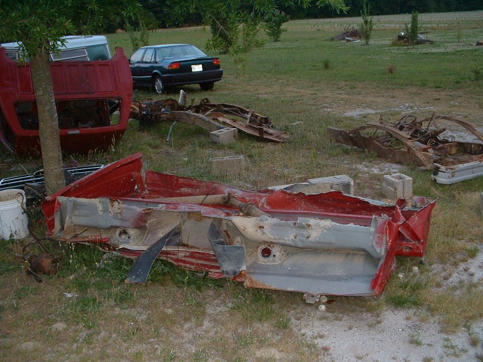 Red truck cab and chassis parts scattered in a yard. A dark sedan is visible in the background