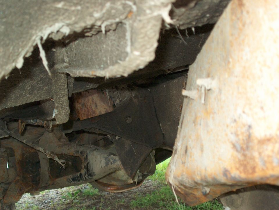 Rusted vehicle underbody with corroded frame and torn insulation over grass