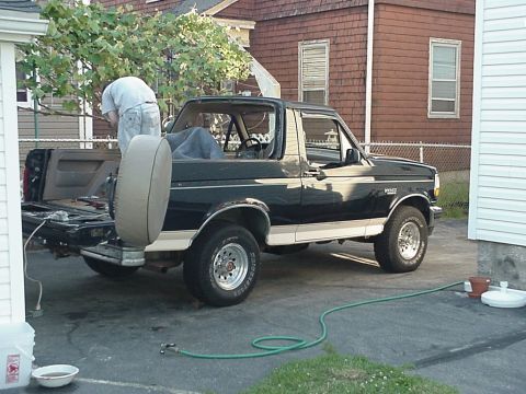 1993 Ford Bronco - A person works on the back of a dark-colored Ford Bronco parked in a driveway next to a house.
