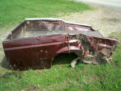 1990 Ford F-150 - This heavily damaged maroon pickup truck bed, likely from a 1990 Ford F-150, sits in a grassy area.