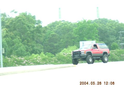 1984 Chevrolet Blazer - A red SUV with large off-road tires is captured in motion, appearing to be airborne. The vehicle's body style suggests a 1995 model year.