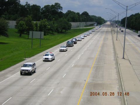 1984 Chevrolet Blazer - A long line of vehicles, including police cars, travels down a highway, likely as part of a funeral procession.