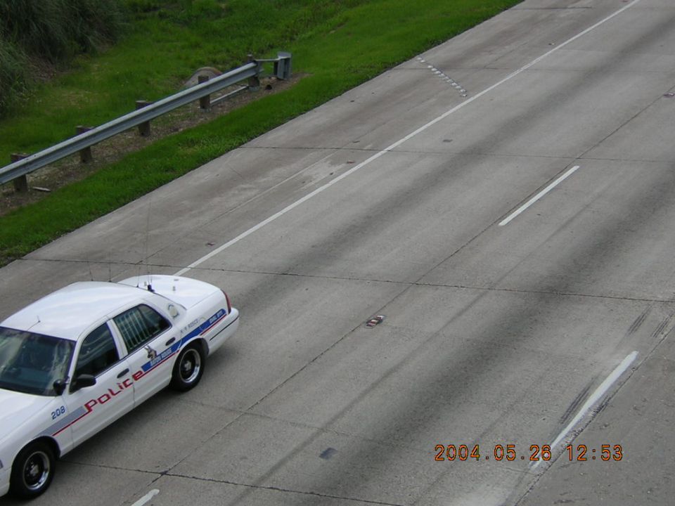 White police car with blue stripe and text 'Police' on its side, seen from above on a highway