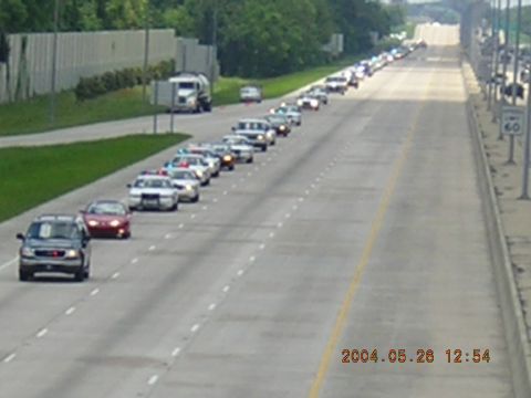 1984 Chevrolet Blazer - A long line of vehicles, including police cars, travels down a highway, likely for a funeral procession.