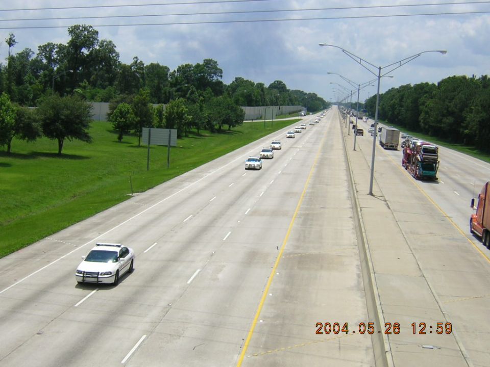 Long line of white cars, possibly police, driving down a highway with trucks on the right