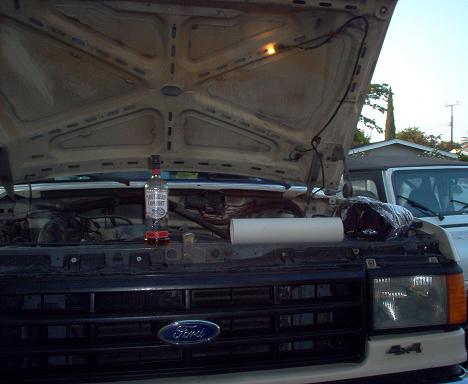 1988 Ford Bronco - The engine bay of a Ford truck is open, with a bottle of Jack Daniels and a roll of paper towels on display. Another vehicle is visible beh…