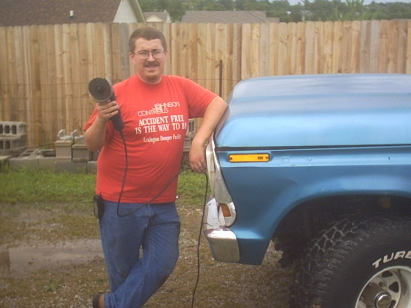 Blue pickup truck fender and headlight with yellow marker light. Man in red shirt leans on it