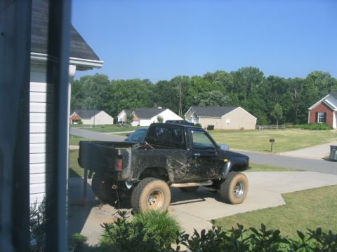 1986 Ford F-150 - A muddy black pickup truck sits on a driveway in front of suburban houses under a clear blue sky.