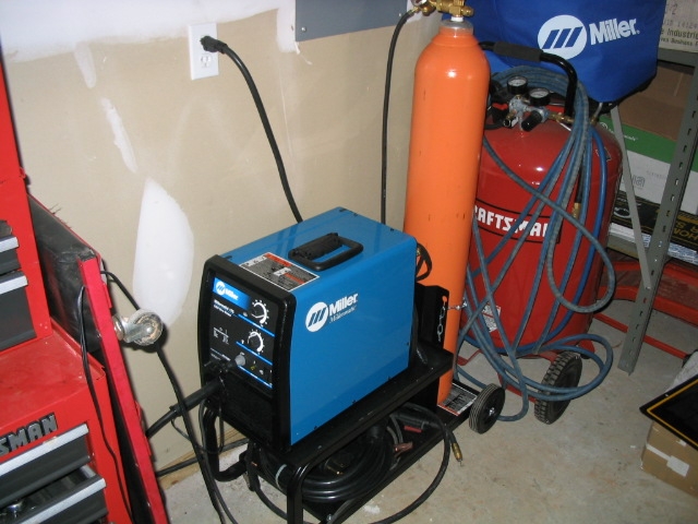 Blue Miller welding machine next to orange gas cylinder and red air compressor