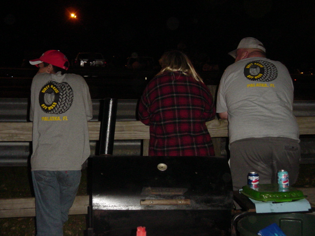 Three people in Palatka, FL tire-logo shirts at a night event behind a black barrel grill