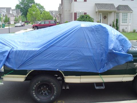 1993 Ford Bronco - A vehicle is covered with a blue tarp, parked in a residential area.