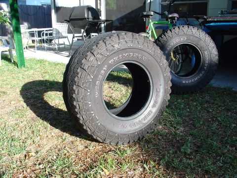 1991 Ford Bronco - Two Mickey Thompson Baja Beltered HP tires are shown in a grassy yard, with a grill and bicycle in the background.