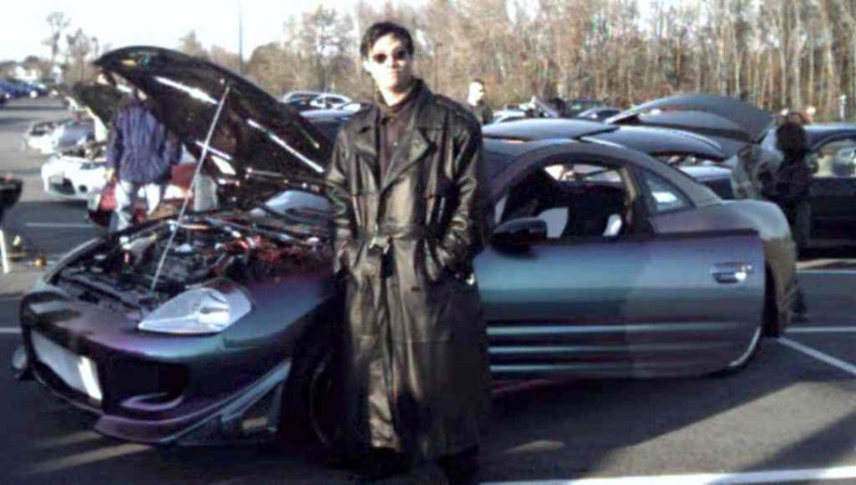 Man in black trench coat next to purple-gray coupe with open hood at a car meet