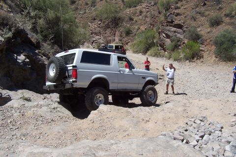 1992 Ford Bronco - A white 1992 Ford Bronco tackles a challenging off-road trail in a desert landscape, with other vehicles and people present.