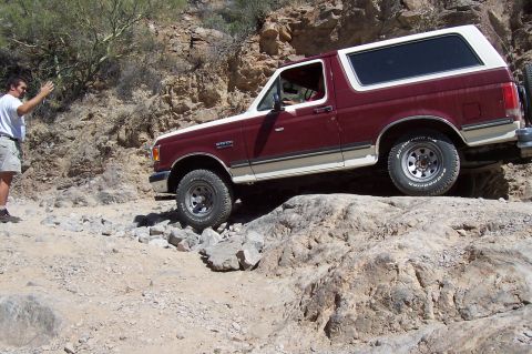 1992 Ford Bronco - 1992 Ford Bronco tackling a challenging rocky off-road path in a desert landscape.