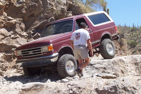1992 Ford Bronco - A man walks next to a maroon Ford Bronco as it climbs a rocky desert path.