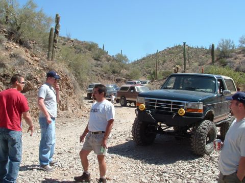 1990 Ford Bronco - Several off-road vehicles are gathered on a dusty trail in a desert landscape with saguaro cacti.