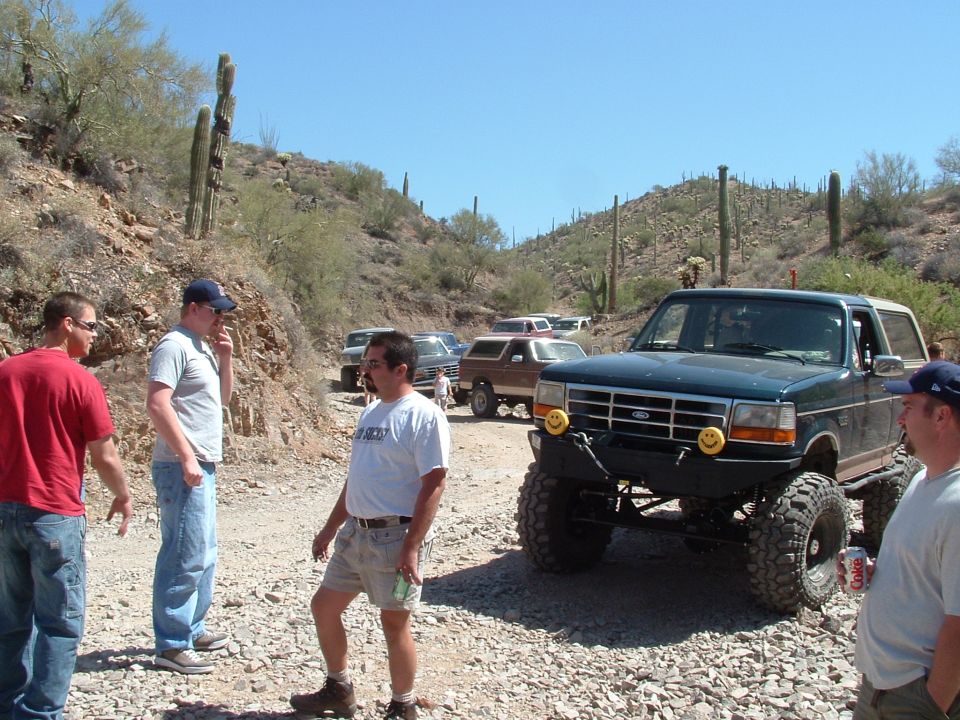Dark green Ford Bronco with large tires and smiley face fog lights parked on a rocky road. Several other SUVs are visible behind it