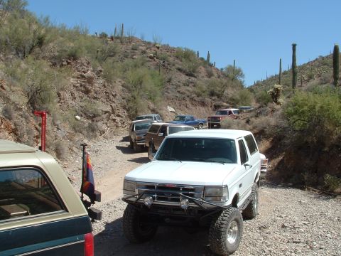 1990 Ford Bronco - A convoy of off-road vehicles navigates a dusty trail through a desert landscape under a clear blue sky.