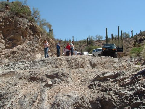 1990 Ford Bronco - A group of people stand near several off-road vehicles on a rocky desert trail, with saguaro cacti in the background.