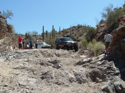 1990 Ford Bronco - Two off-road vehicles navigate a challenging rocky trail in a desert canyon with saguaro cacti.