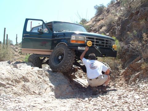 1990 Ford Bronco - A man adjusts a smiley face light on the bumper of a lifted Ford truck navigating a rocky desert trail.