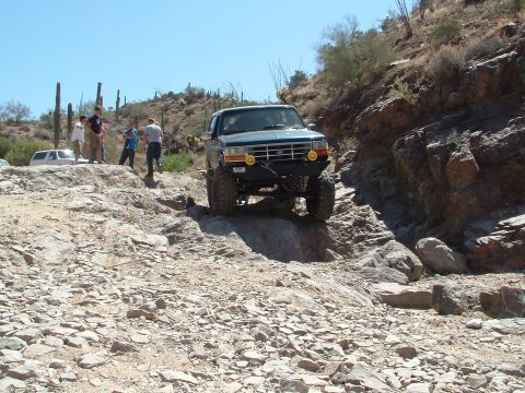 1990 Ford Bronco - A lifted Ford Bronco navigates a challenging rocky trail in a desert landscape with cacti.