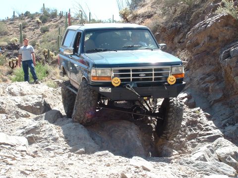 1990 Ford Bronco - A teal Ford Bronco drives over rocks on a desert trail.
