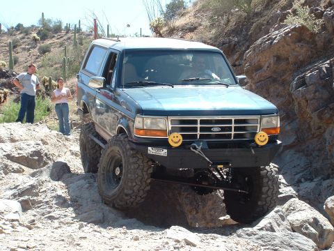 1990 Ford Bronco - This blue SUV is shown off-roading on a rocky desert trail with two people standing nearby.