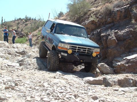 1990 Ford Bronco - 1990 Ford Bronco tackling a challenging rocky trail with desert vegetation in the background.