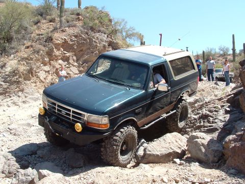 1990 Ford Bronco - A modified Ford Bronco tackles a challenging off-road course with other vehicles and people present.