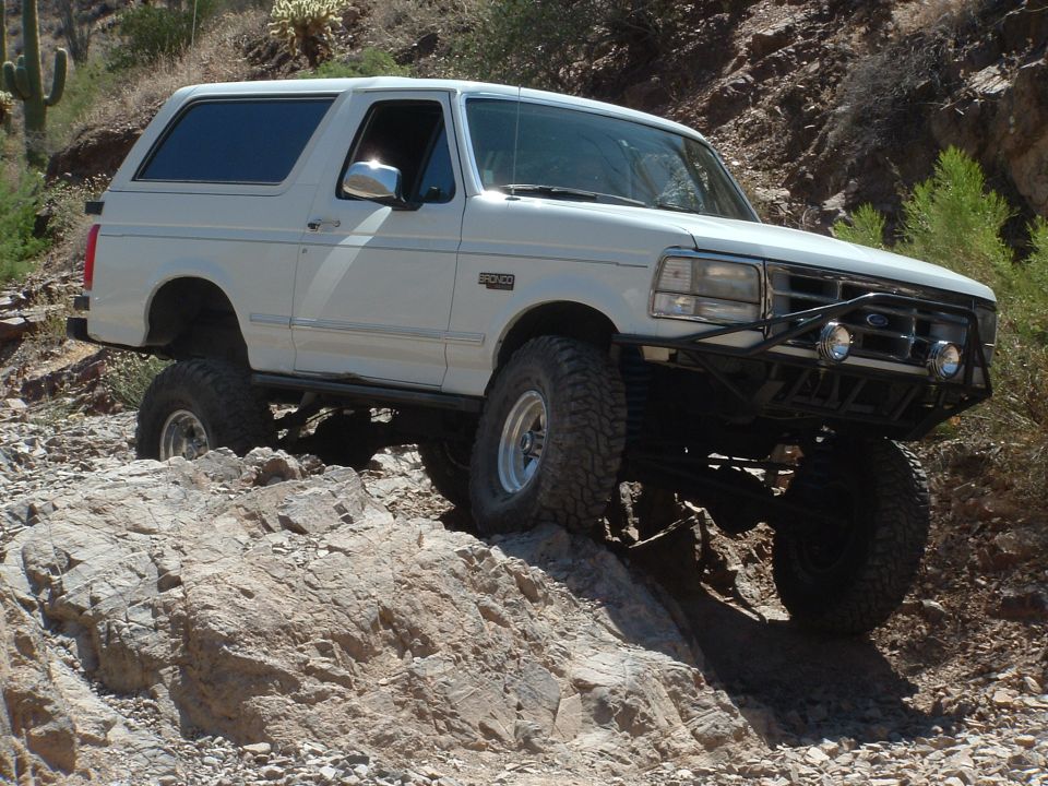 White 1990 Ford Bronco with off-road tires and bumper driving over rocks