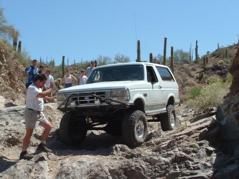 1990 Ford Bronco - A white Ford Bronco is driven over a rocky desert path with several people watching.