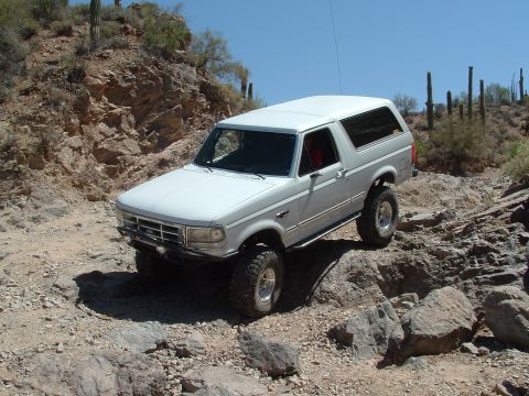 1990 Ford Bronco - A white Ford Bronco navigates a rocky desert terrain under a clear blue sky.