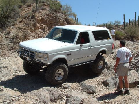 1990 Ford Bronco - A white Ford Bronco navigates a rocky desert terrain with a person standing nearby.