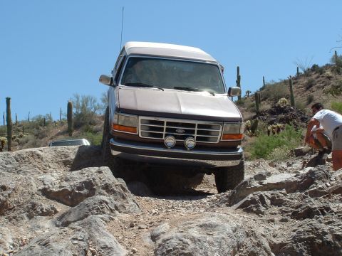 1990 Ford Bronco - A brown Ford Bronco drives over rocks in a desert landscape with cacti.