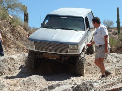 1990 Ford Bronco - A man walks alongside a silver SUV as it navigates a challenging rocky trail in a desert environment.